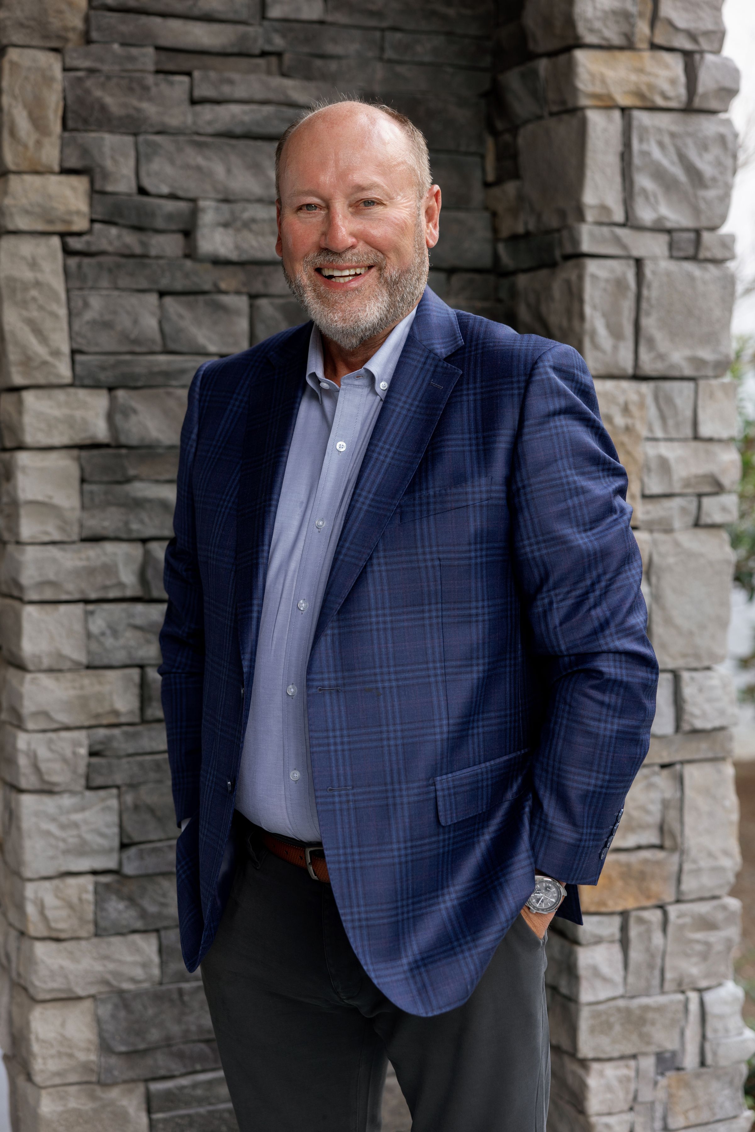 Professional business portrait of someone in a navy plaid blazer and gray shirt standing against a stone wall backdrop.