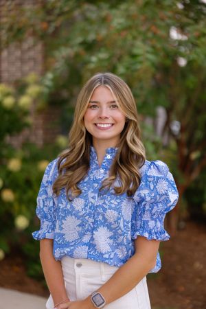 A person in a blue and white floral patterned blouse with puff sleeves smiles in an outdoor setting.