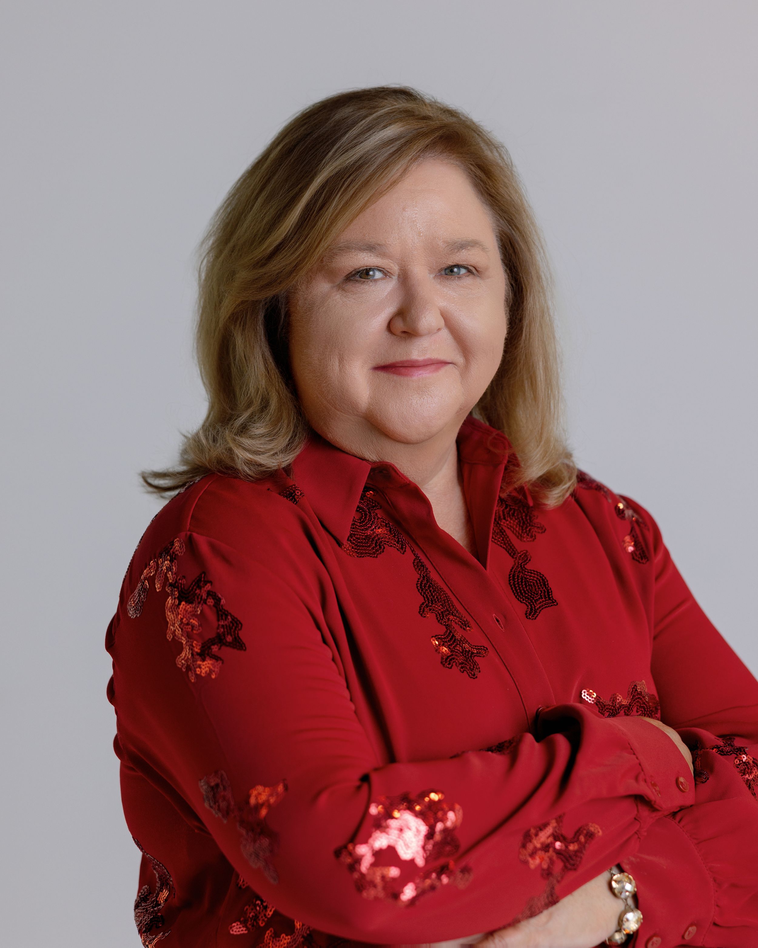 A person wearing a festive red holiday sweater decorated with sequins smiles warmly in front of a gray background.