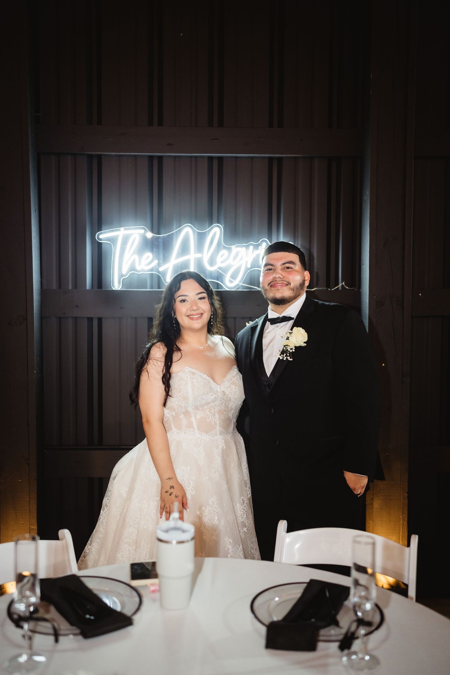 A couple in formal wedding attire stands at their reception table with a neon sign illuminated behind them.