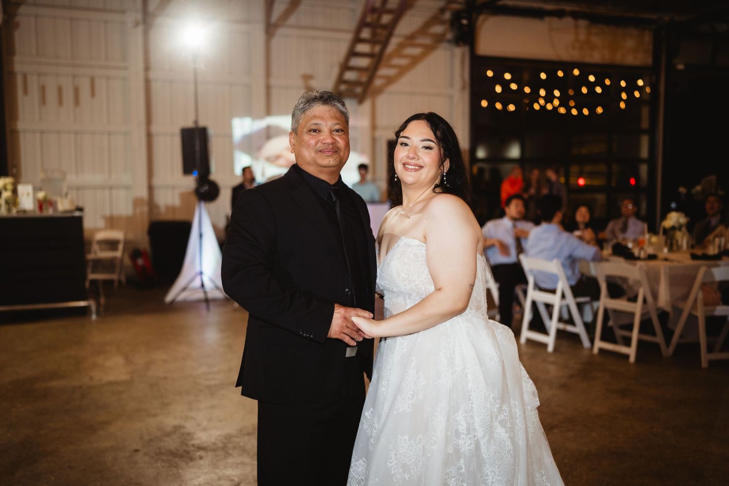 A couple shares a romantic dance together at an indoor venue decorated with twinkling lights and white chairs.