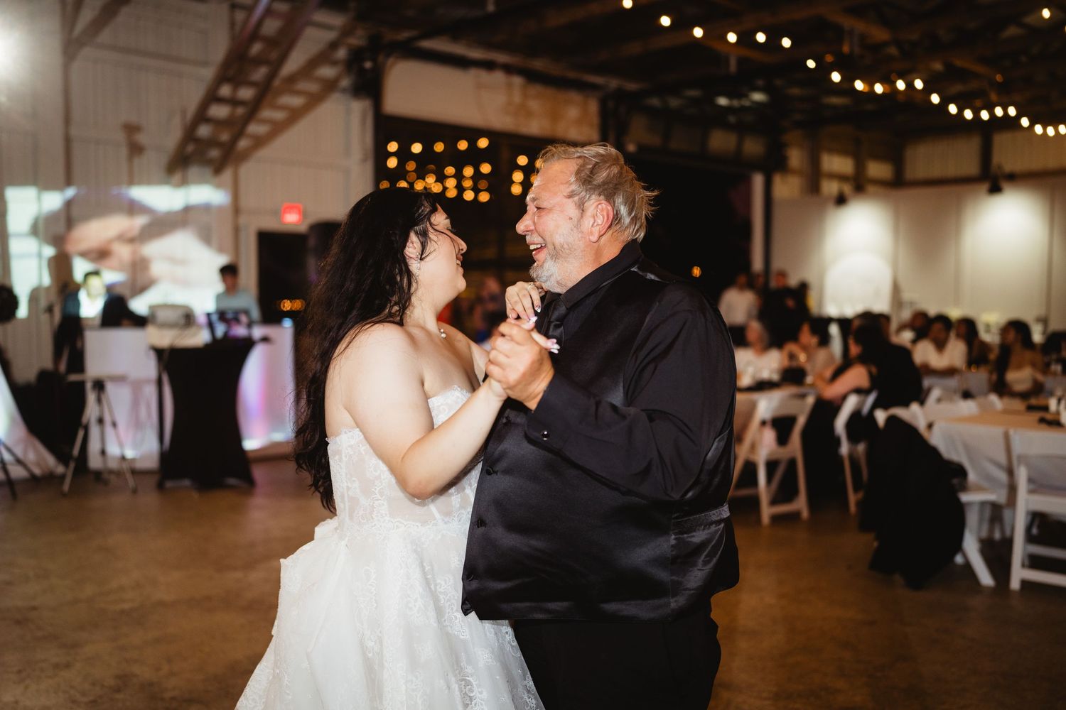 Couple shares a special dance moment at a reception venue decorated with string lights and featuring wooden chairs.