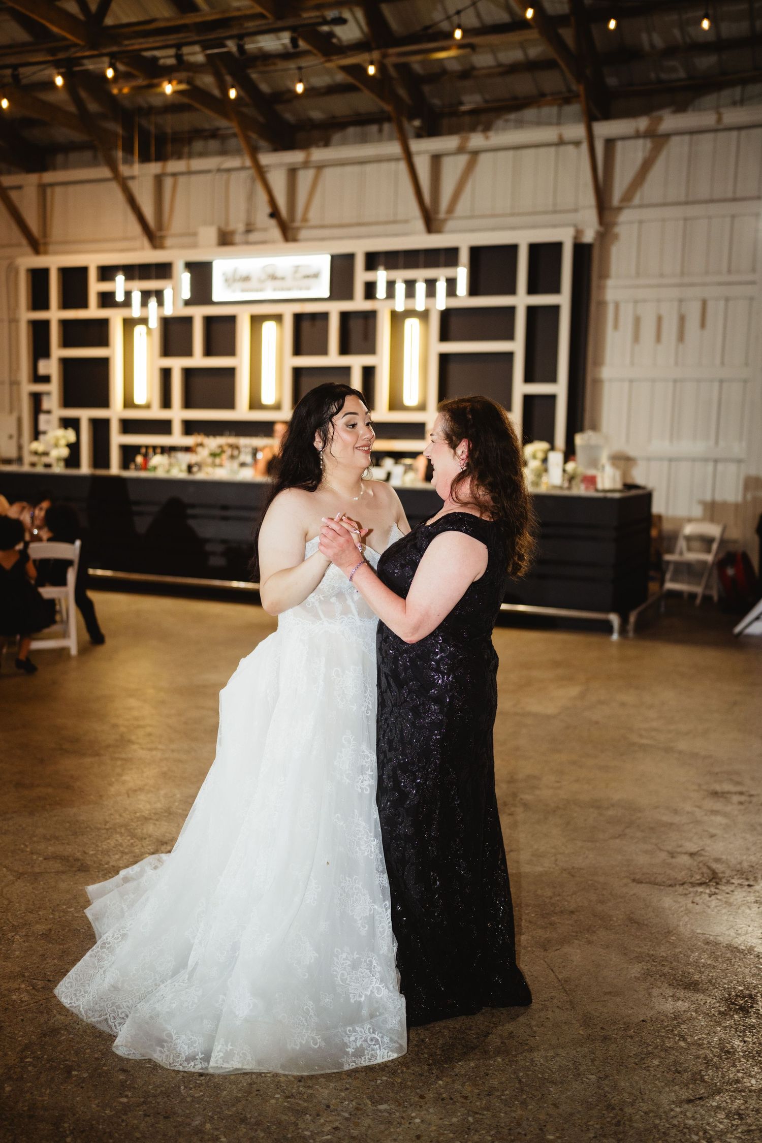 Two people in formal attire dance together in a rustic venue with string lights and a bar in the background.