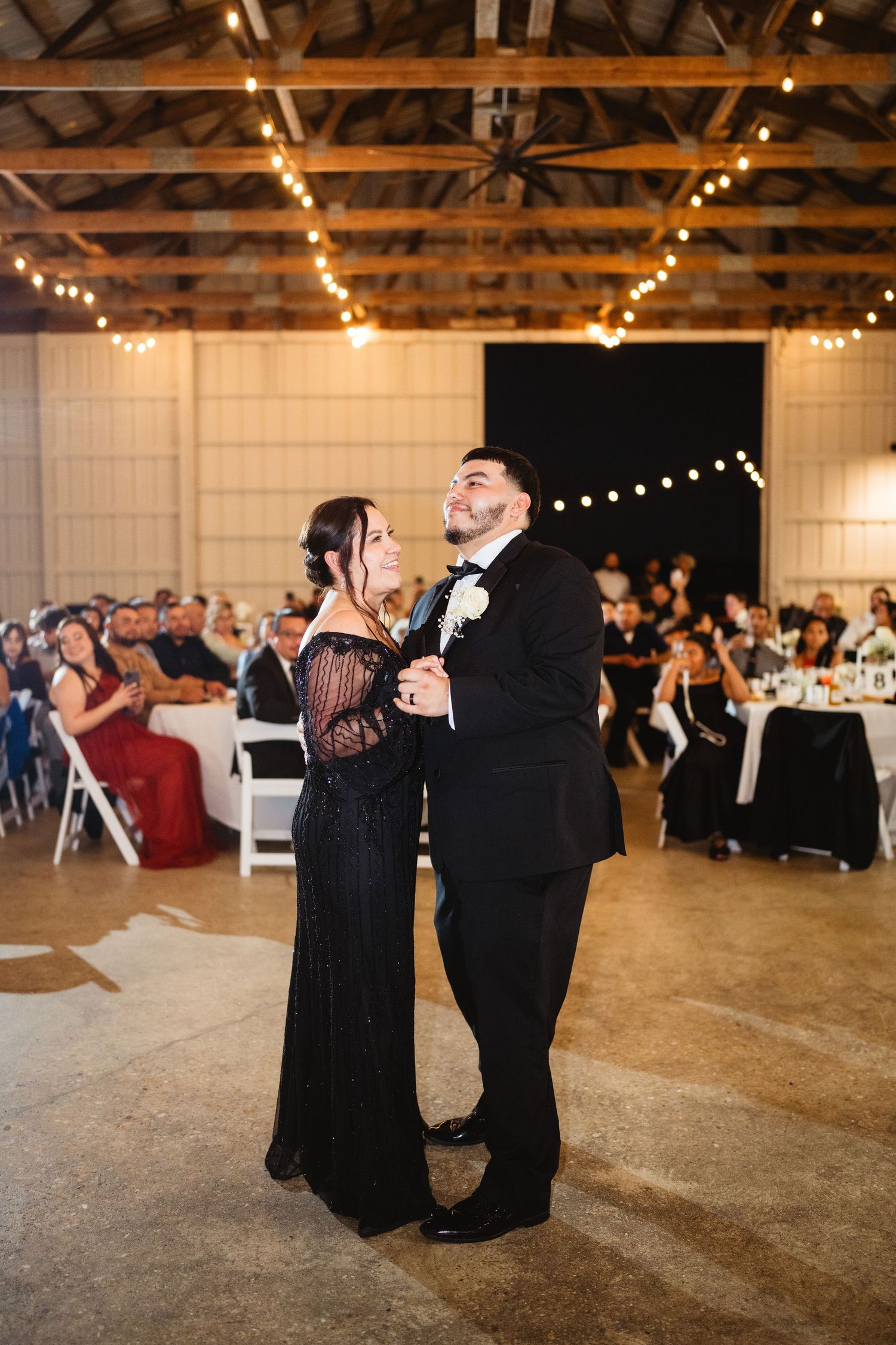 Wedding guests dance together under string lights in a rustic barn venue at an evening reception.