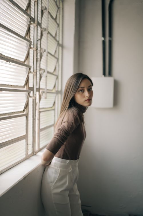 A moody indoor portrait of someone in a brown turtleneck and white pants standing by a window with venetian blinds.
