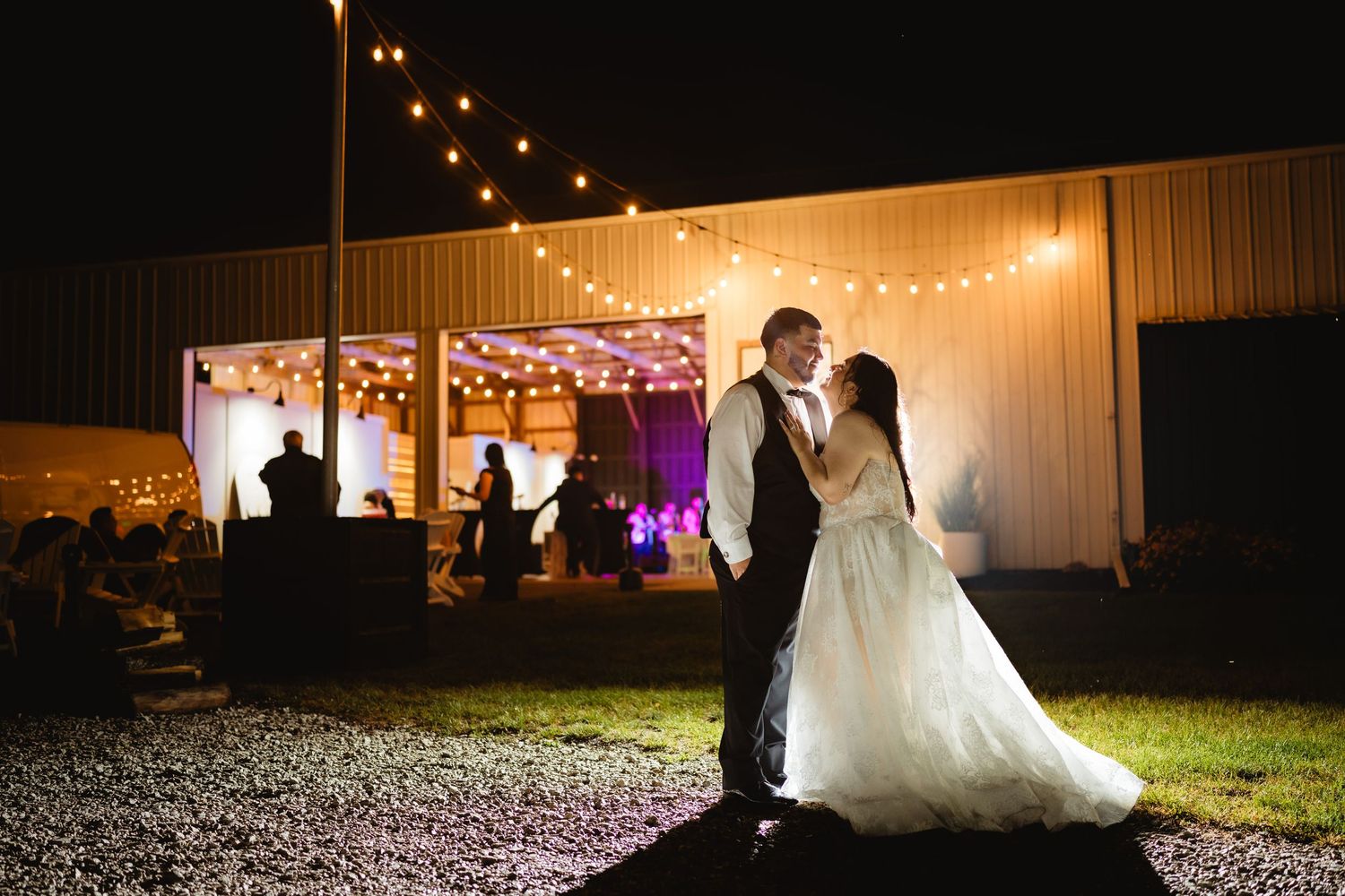 Beautiful evening wedding photos captured at a rustic white barn venue illuminated by warm outdoor lighting at night.