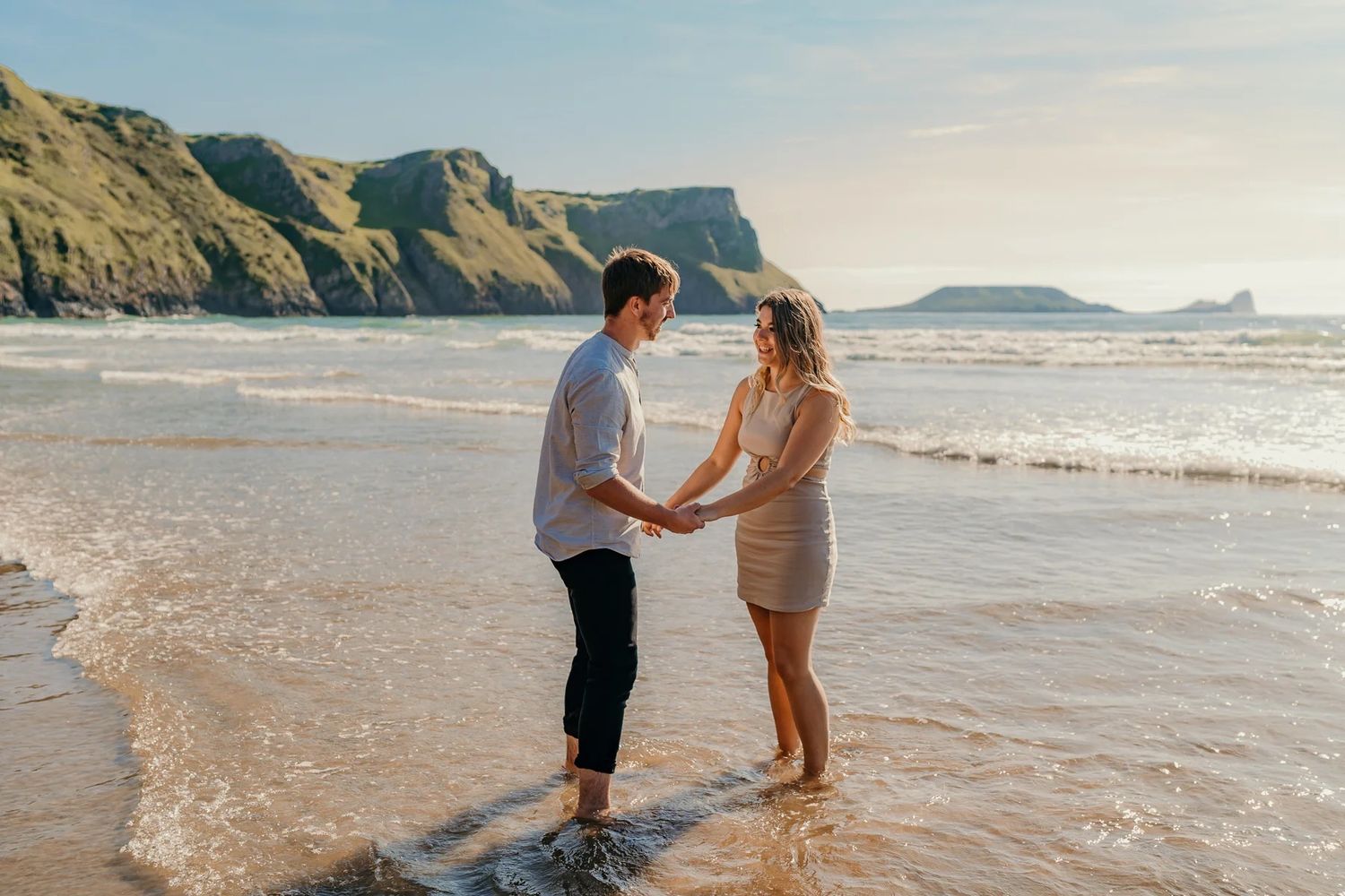 Couple playfully splashing and running through ocean waves along a scenic rocky coastline at sunset.