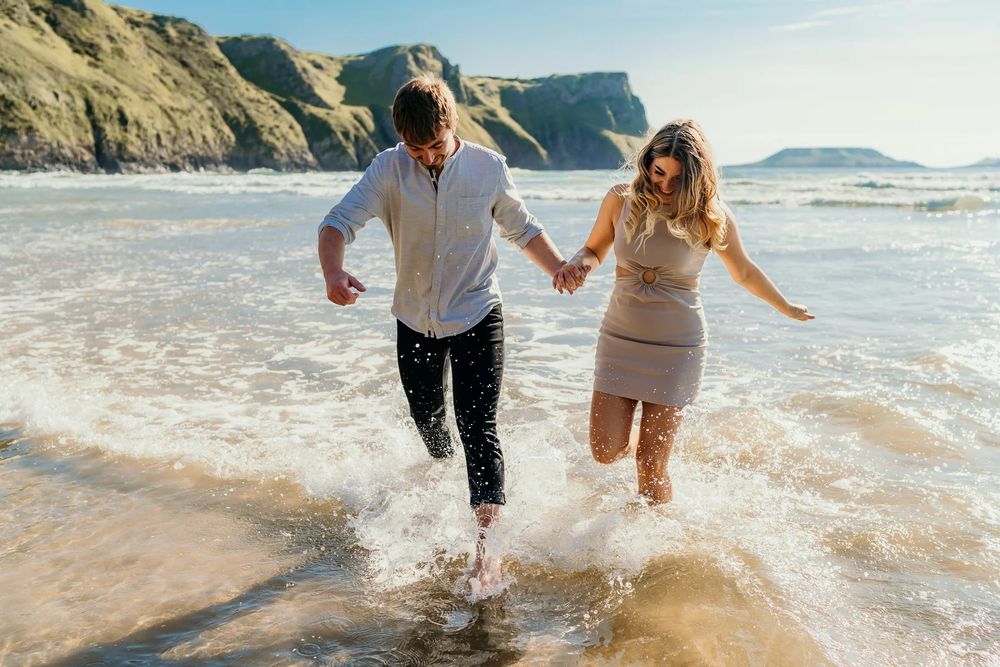 Couple enjoying a romantic moment while walking barefoot through shallow ocean waves near coastal cliffs.