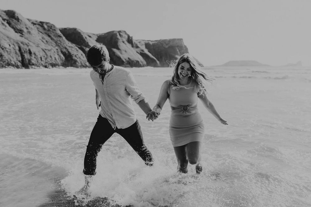 Black and white photo of couple playing and laughing together in ocean surf near dramatic cliffs.