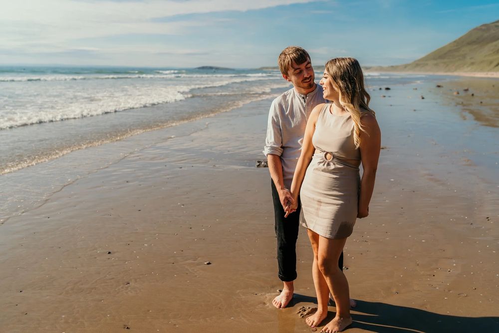 A couple walking together at sunset along a scenic coastal beach with rocky cliffs in the background.