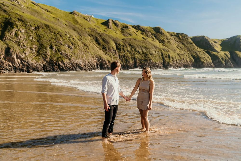 People walk along a sandy beach surrounded by rugged cliffs on a sunny day at the coast.