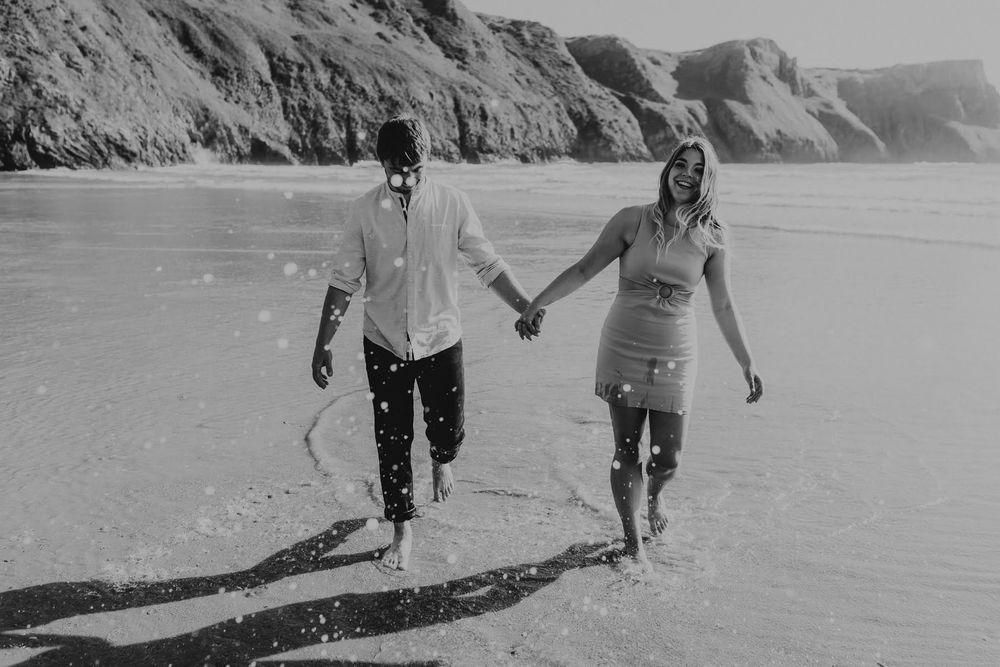 Black and white shot of a couple holding hands and running joyfully on a sandy beach by rugged cliffs.