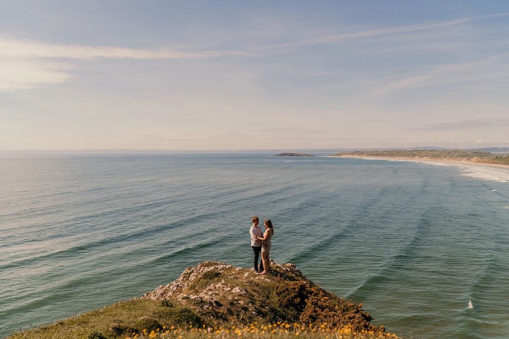 Coastal panoramic view of dramatic cliffs overlooking a calm blue ocean with figures silhouetted against the horizon.