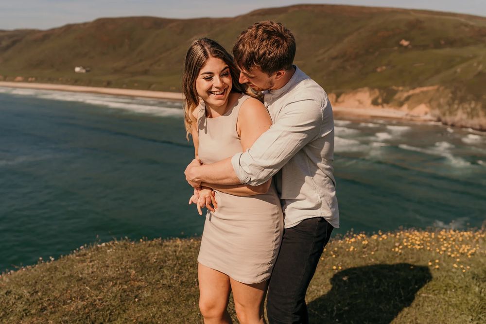 A couple embraces on a scenic coastal cliff overlooking the ocean during golden hour.