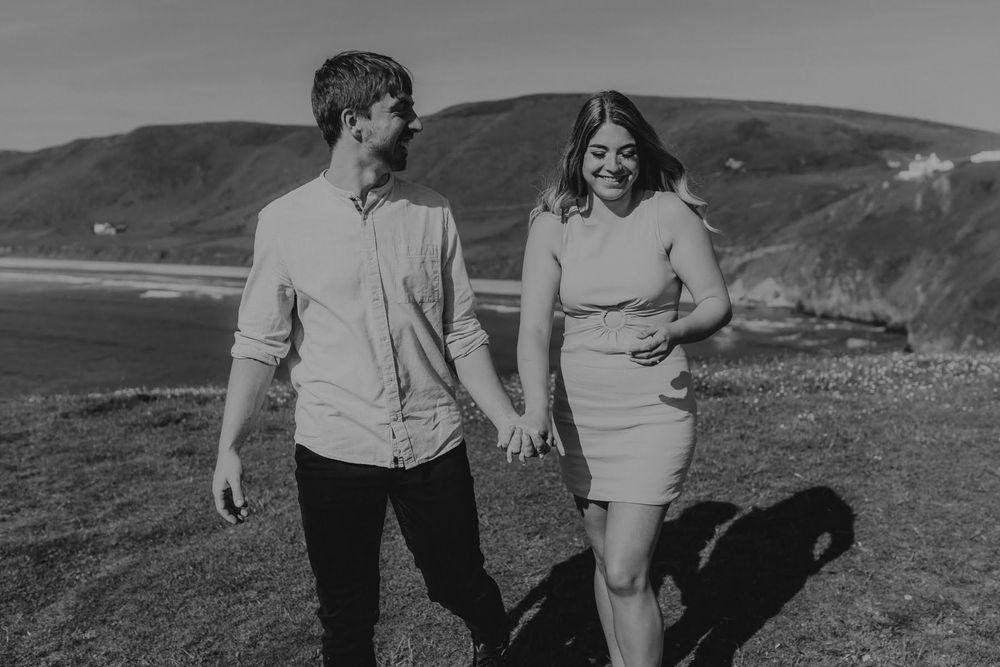 Couple walks hand in hand along scenic coastal cliffs on a sunny afternoon in black and white.