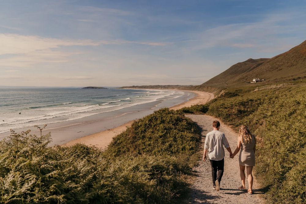 Couple on coastal path overlooking sandy beach at sunset during romantic seaside walk along grassy cliffs.