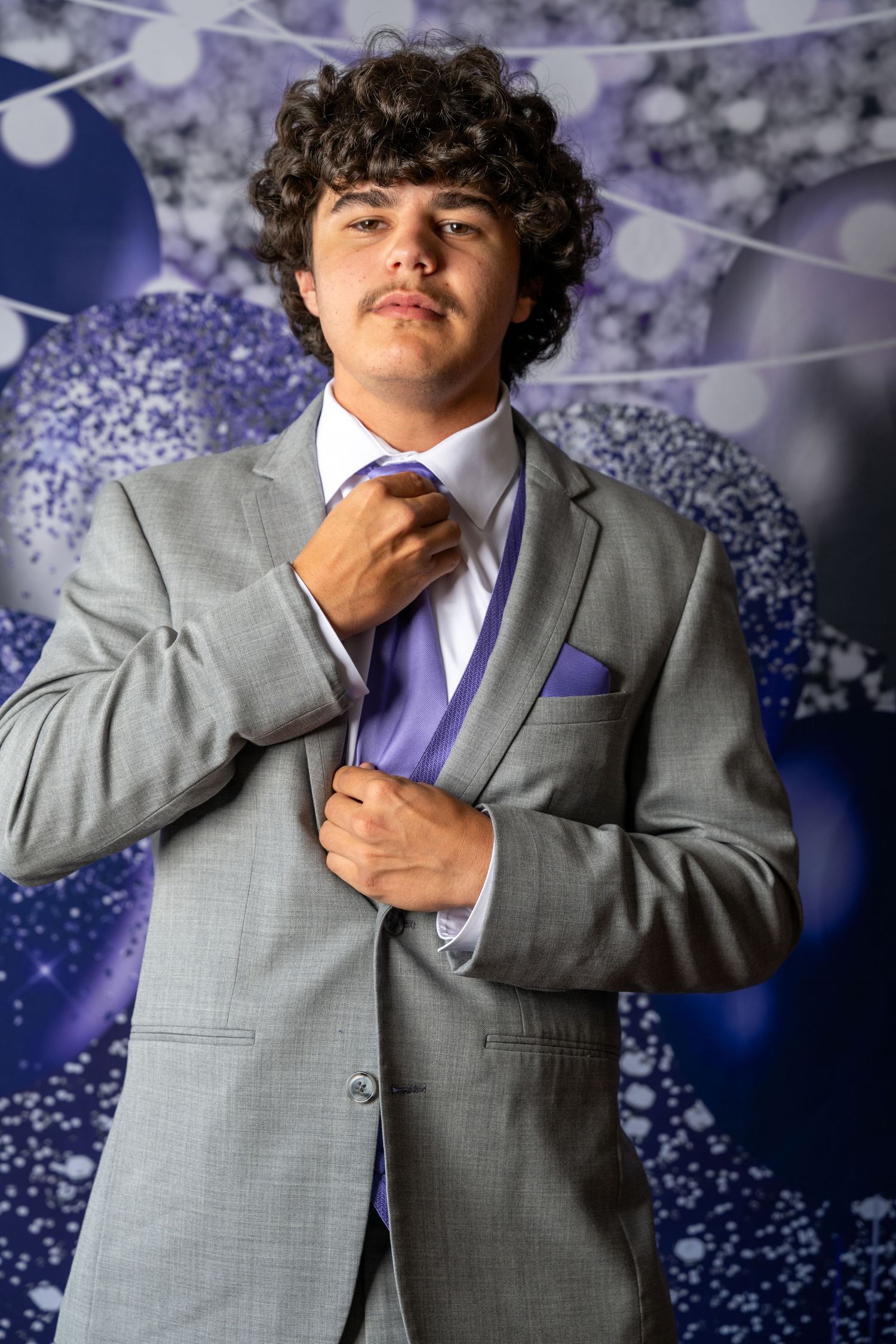 Formal portrait of someone in a light gray suit with purple tie adjusting their collar against a sparkly blue backdrop.