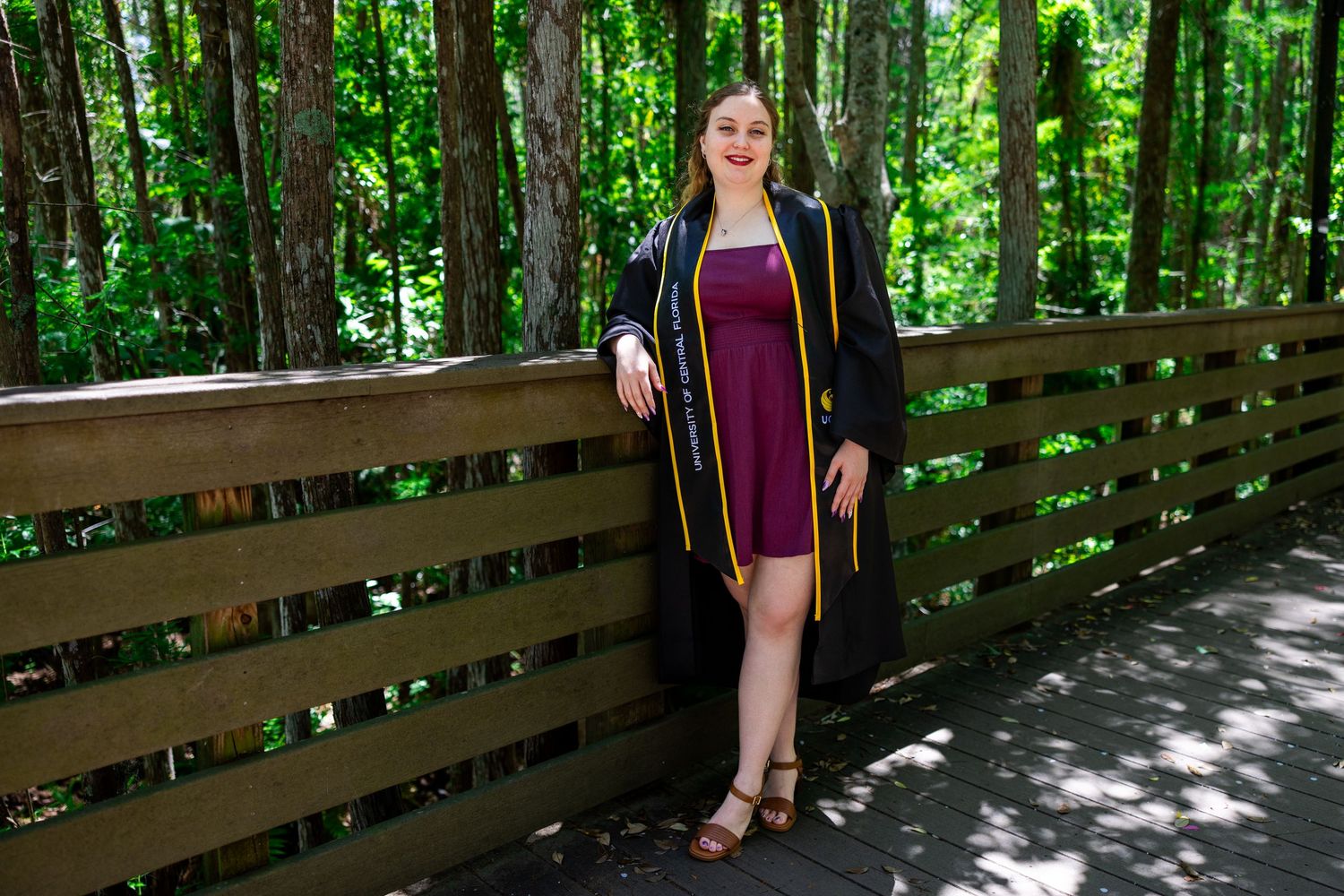 A person in a burgundy dress and black jacket stands on a sunlit wooden boardwalk surrounded by lush green forest.