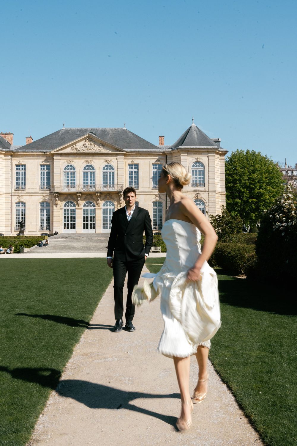 A couple poses on the lawn in front of an elegant French chateau with white stone facade and classical architecture.