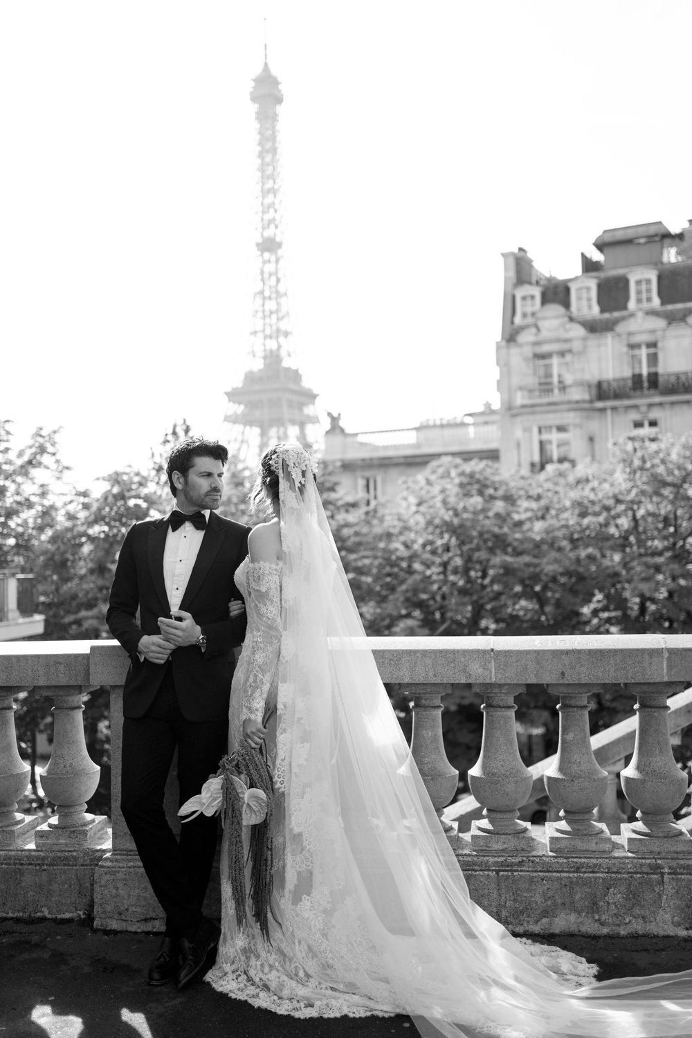 Bride and groom in formal attire share a romantic moment on an elegant stone balcony with European architecture in the background.