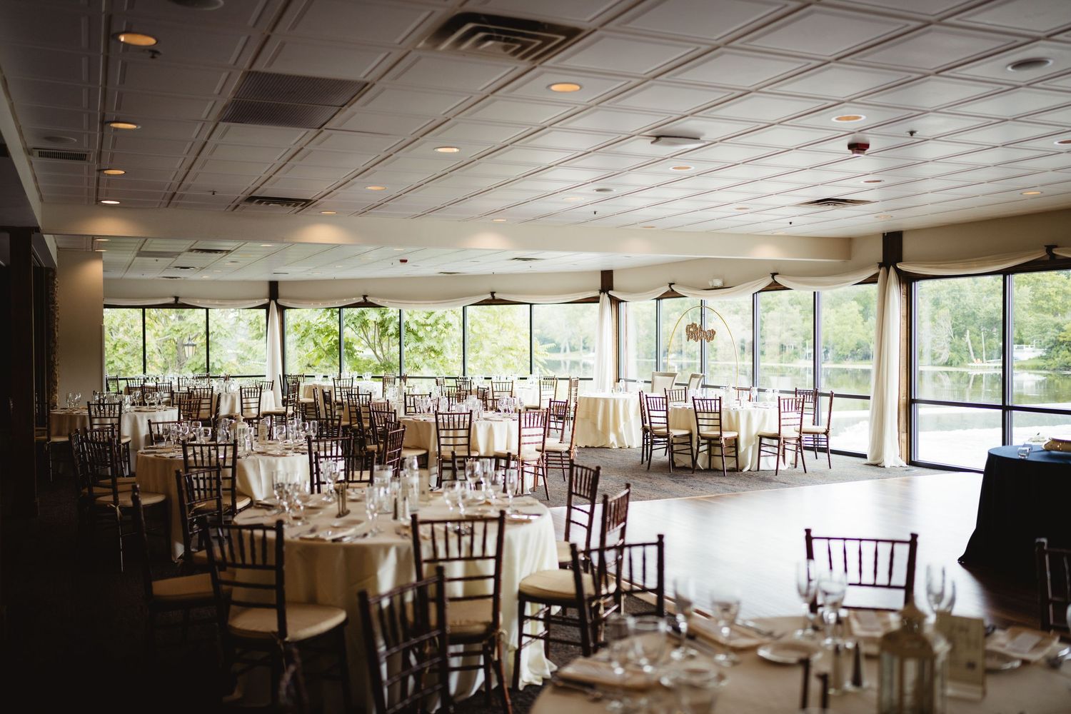 Event venue interior with wooden chiavari chairs set up around dining tables in an open ballroom with scenic window views.