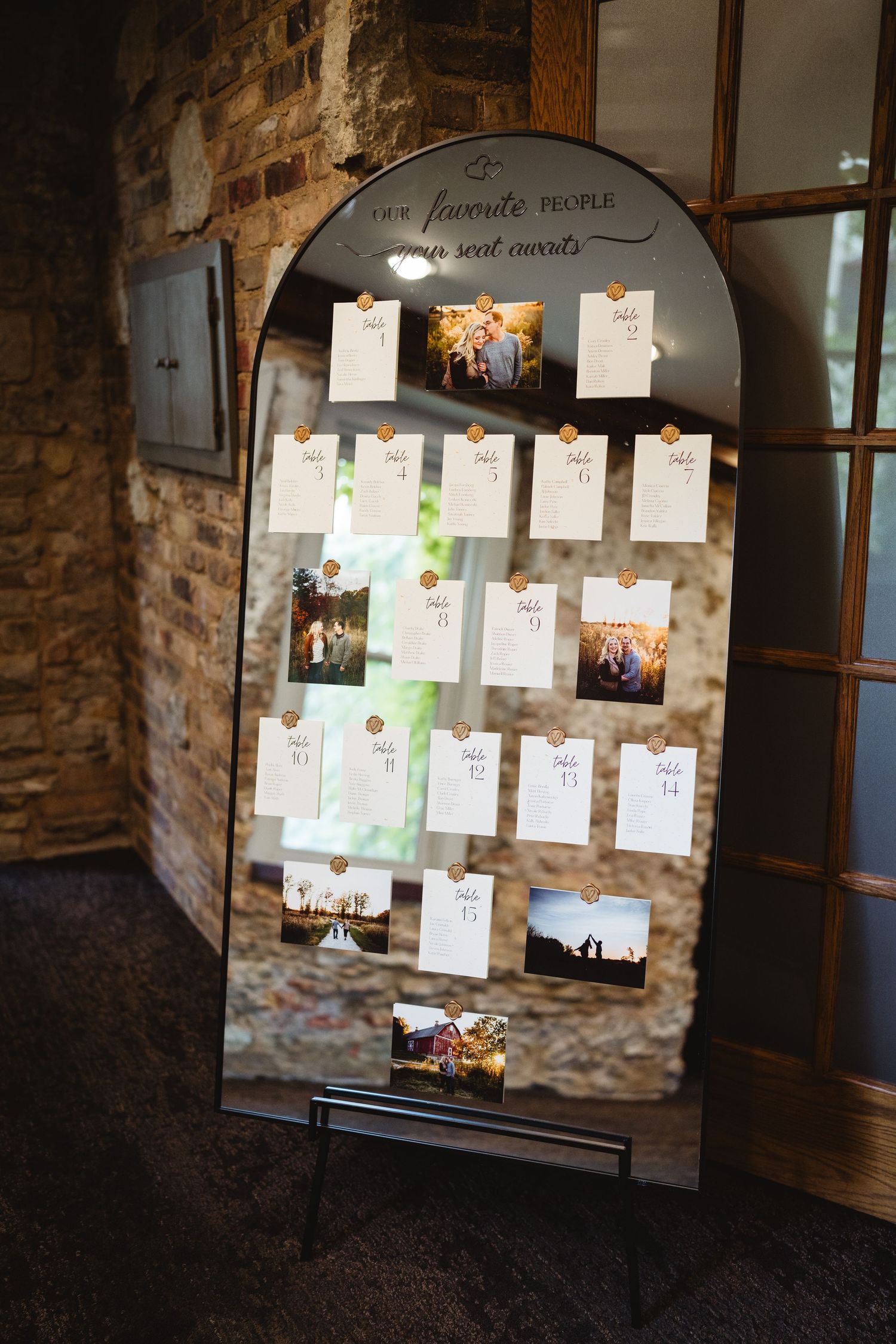 A vintage arched mirror displays wedding photos and seating cards arranged on strings against exposed brick wall decor.