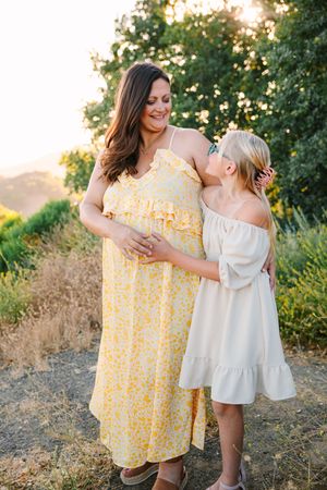 A tender moment captured at sunset with a mother and daughter in white and yellow floral dresses among summer foliage.