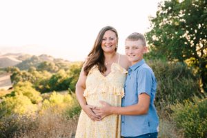 Mother in yellow dress and boy in blue shirt pose together at sunset on a hillside with golden light.