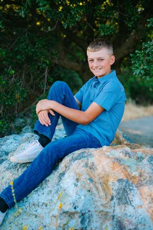 A young person in a blue polo shirt and jeans sits casually on a large rock outdoors surrounded by green foliage.