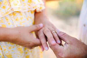 Two hands wearing wedding rings gently touch in a soft, romantic moment outdoors with sunlit bokeh in background.