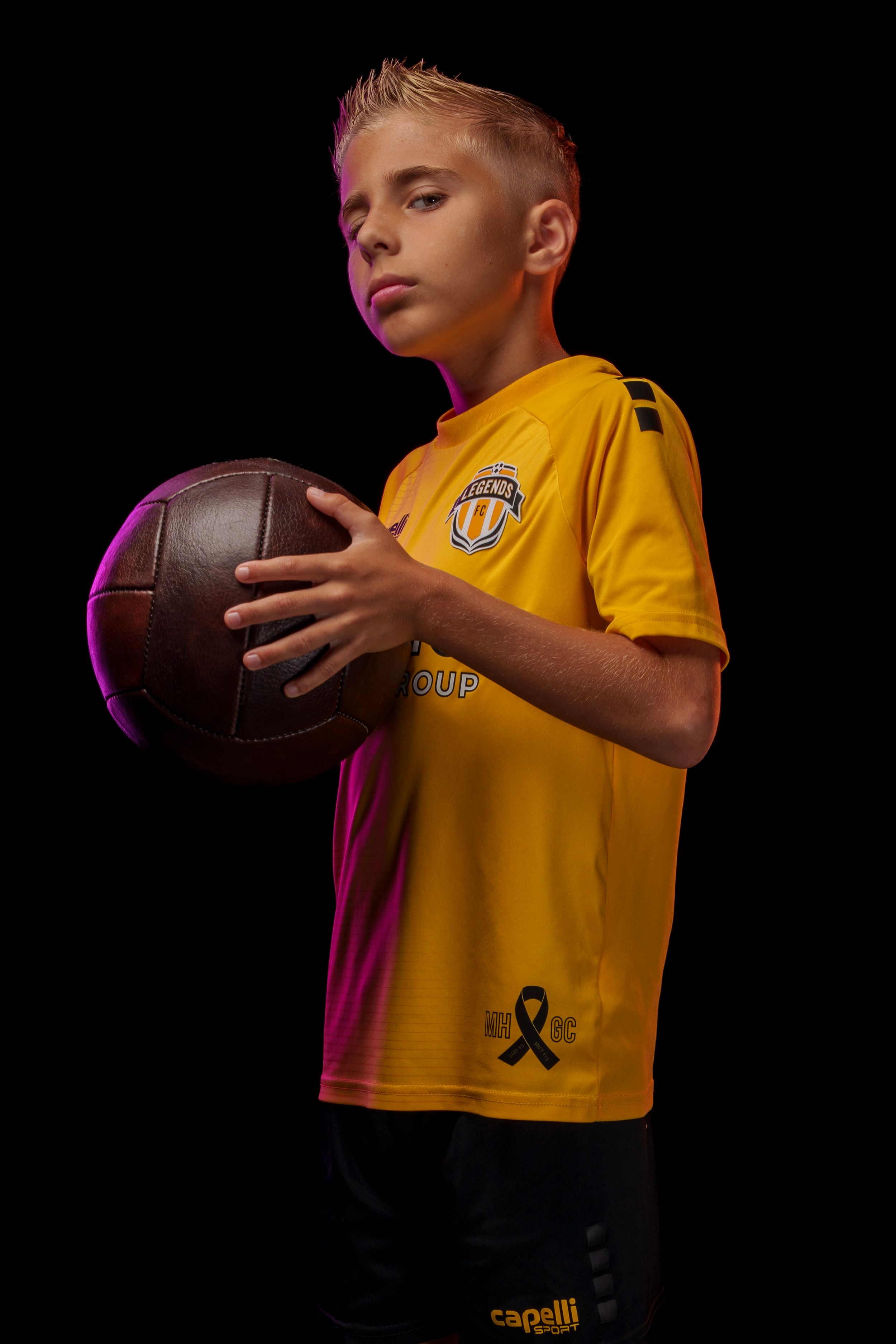 Youth athlete poses with soccer ball against dark background in yellow Legends FC  jersey.