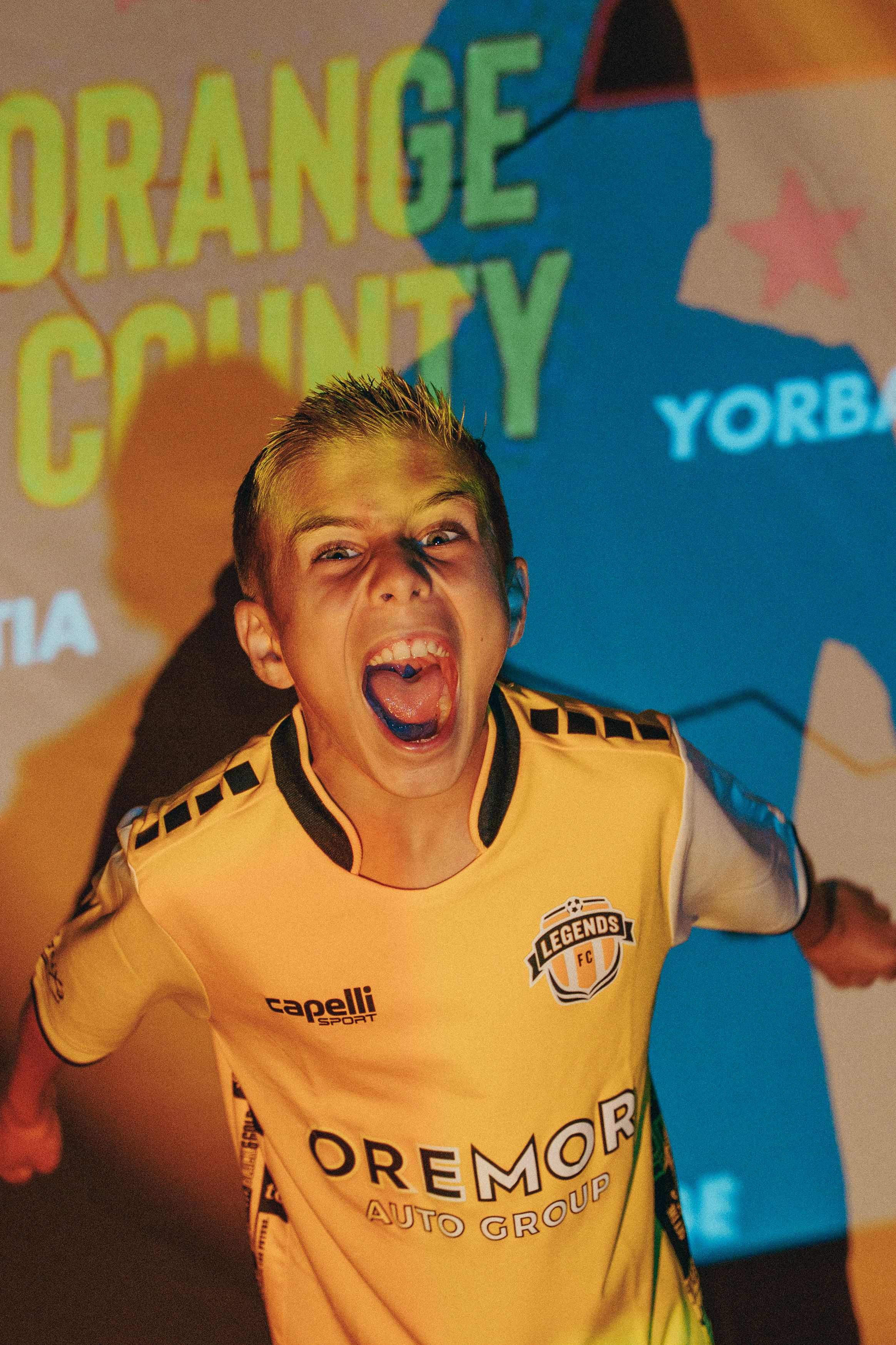 Energetic poses in yellow soccer uniform against orange and blue backdrop.