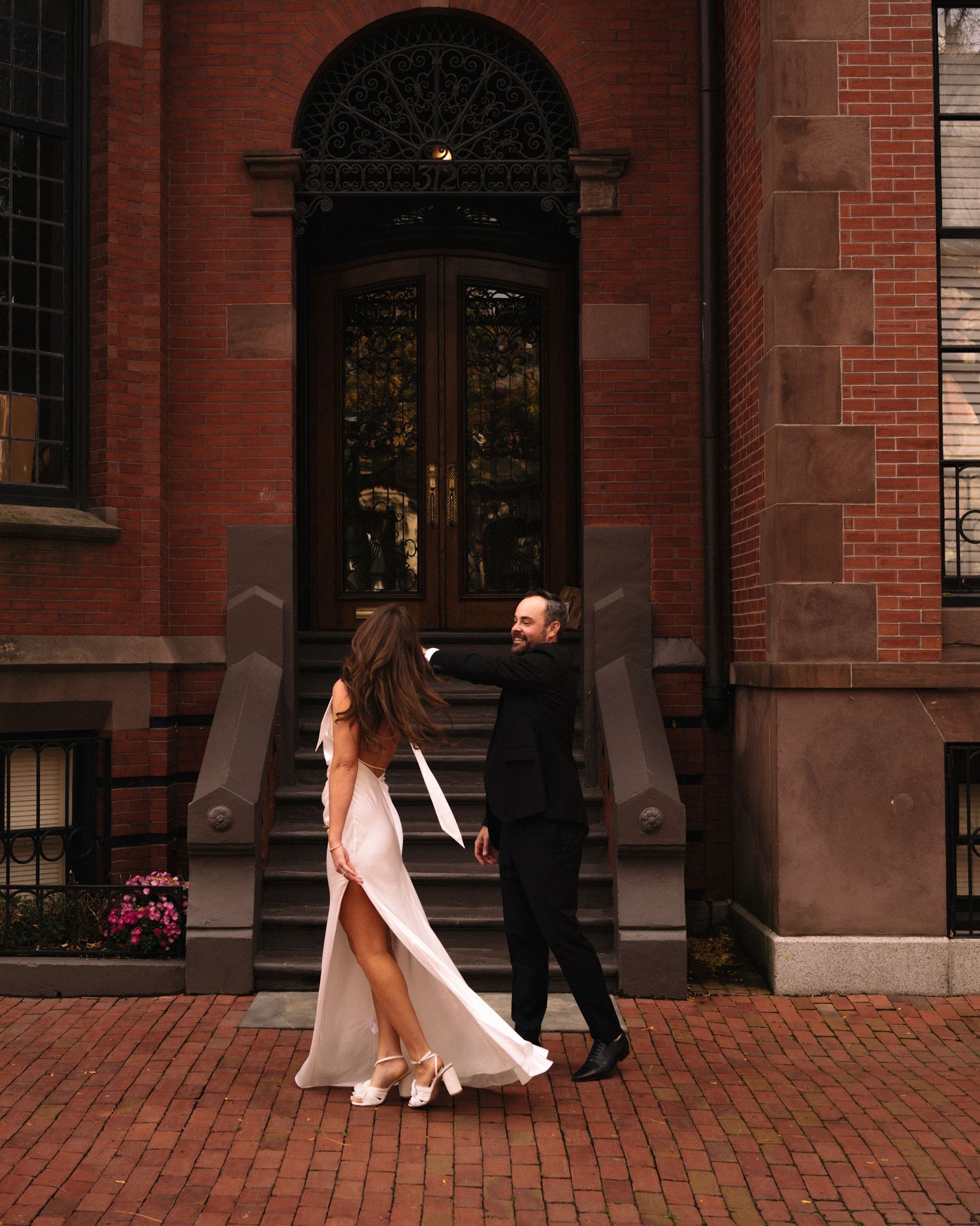 Couple in formal wear walking along a red brick building with arched doorways and windows at sunset.
