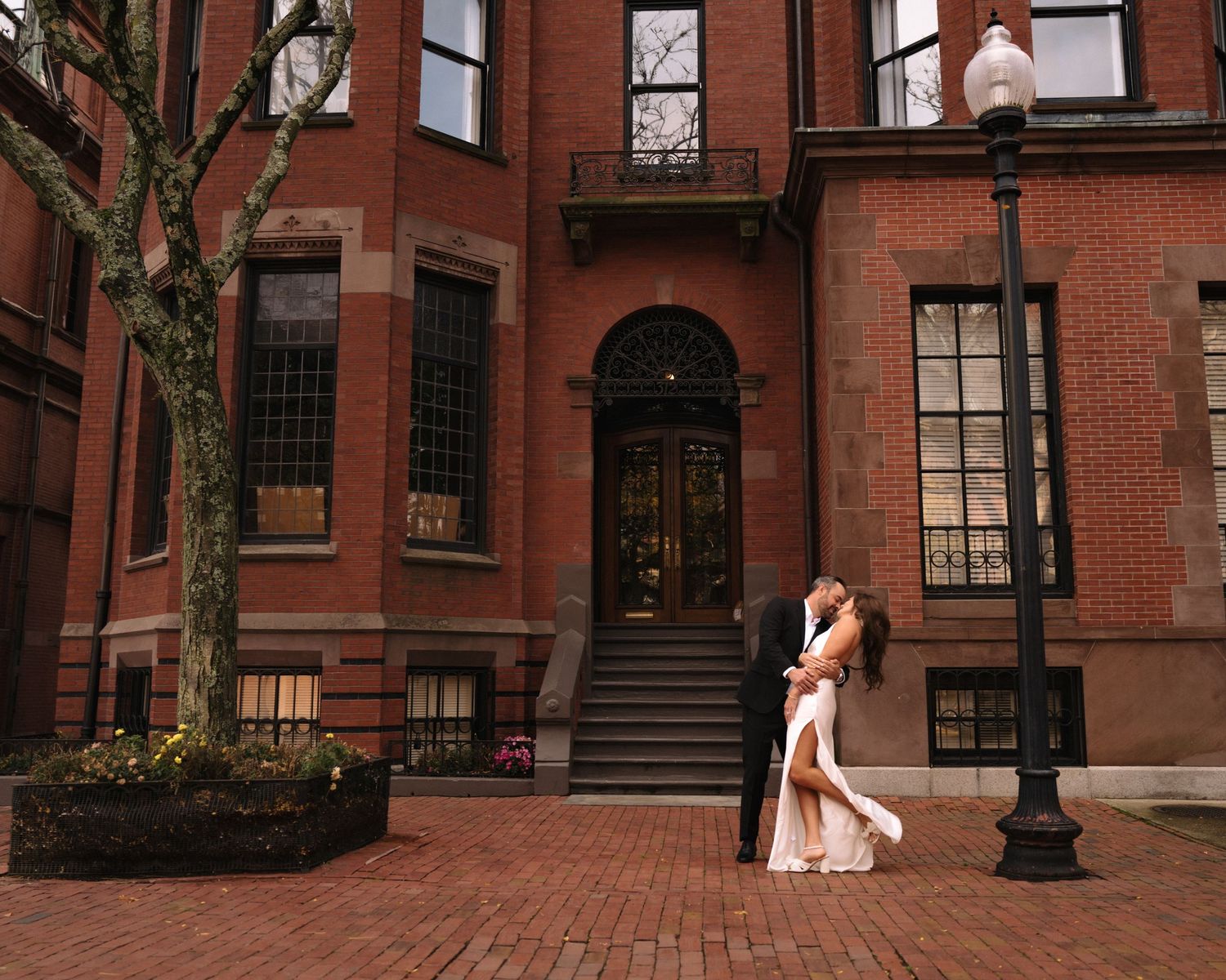Elegant couple poses on red brick sidewalk in historic Boston neighborhood with brownstone buildings.