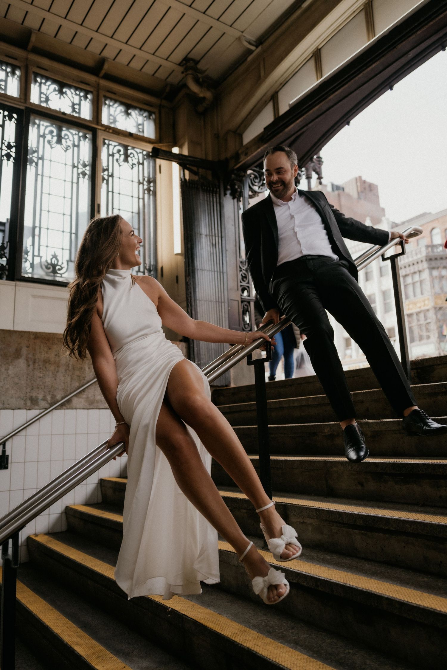 A couple on wooden stairs at a subway station playfully dance and pose together in wedding attire.