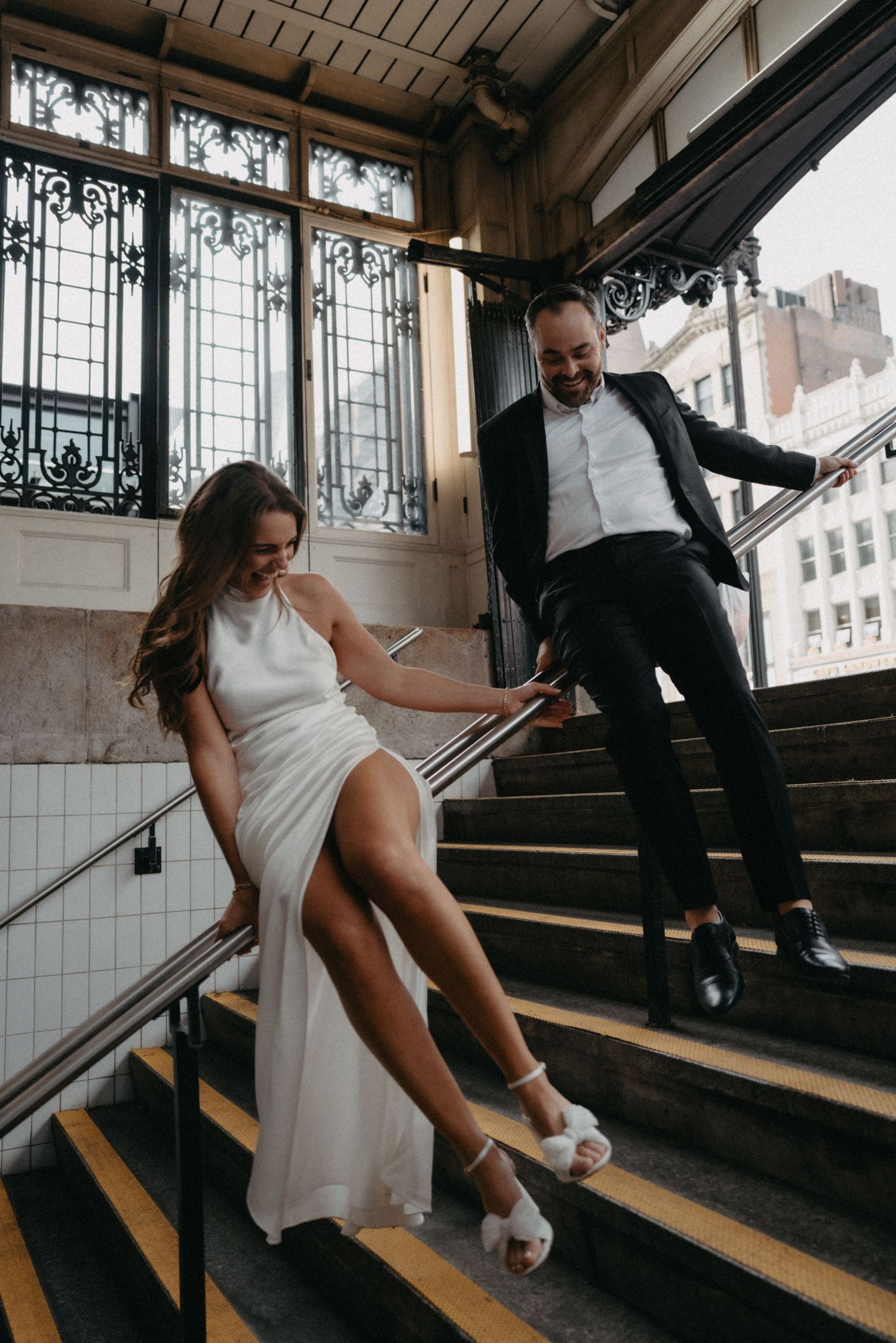 A couple in formal attire pose playfully on an elegant vintage staircase with ornate iron railings and window details.
