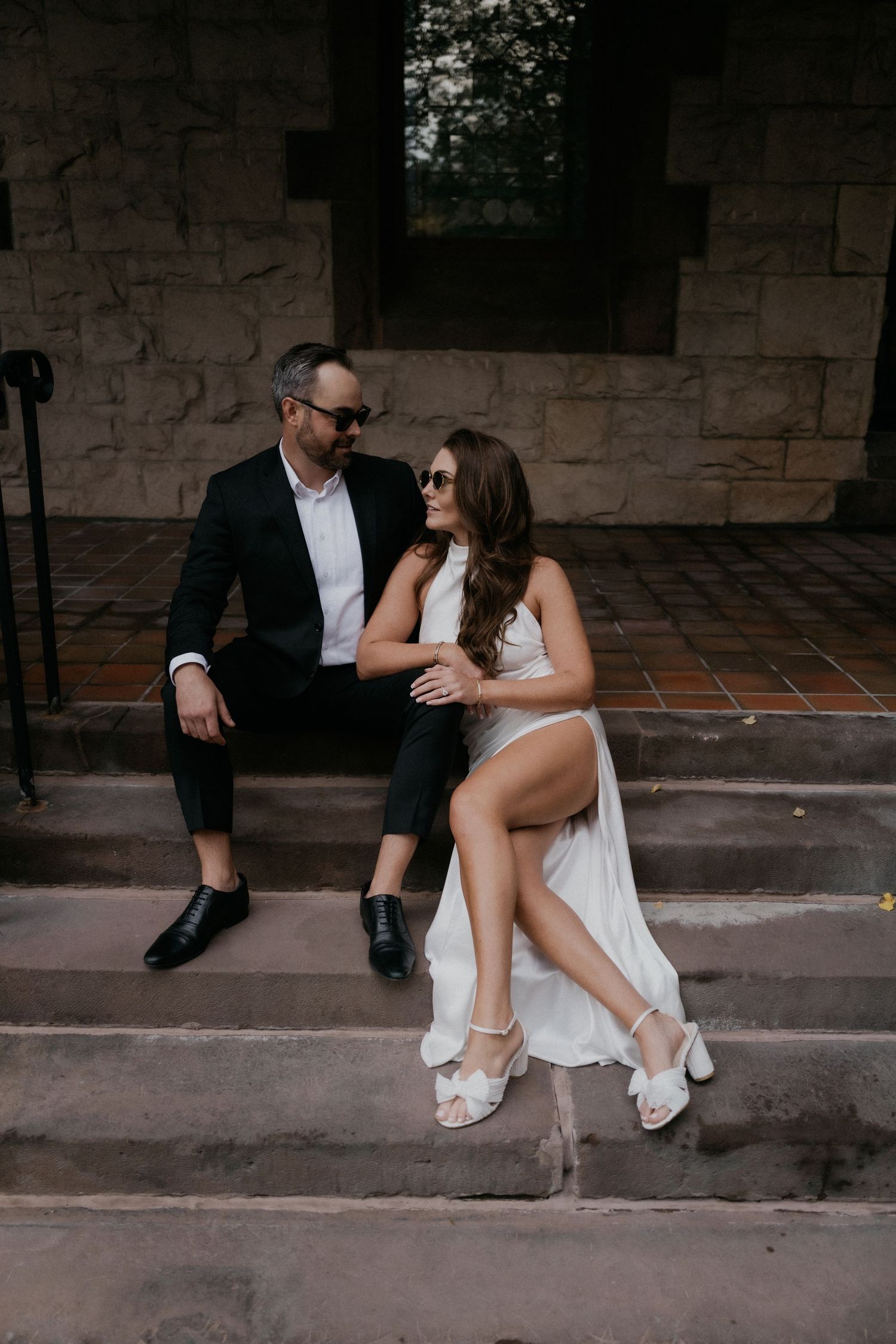 A couple in wedding attire sits intimately on stone steps, sharing a romantic moment in their white and black outfits.