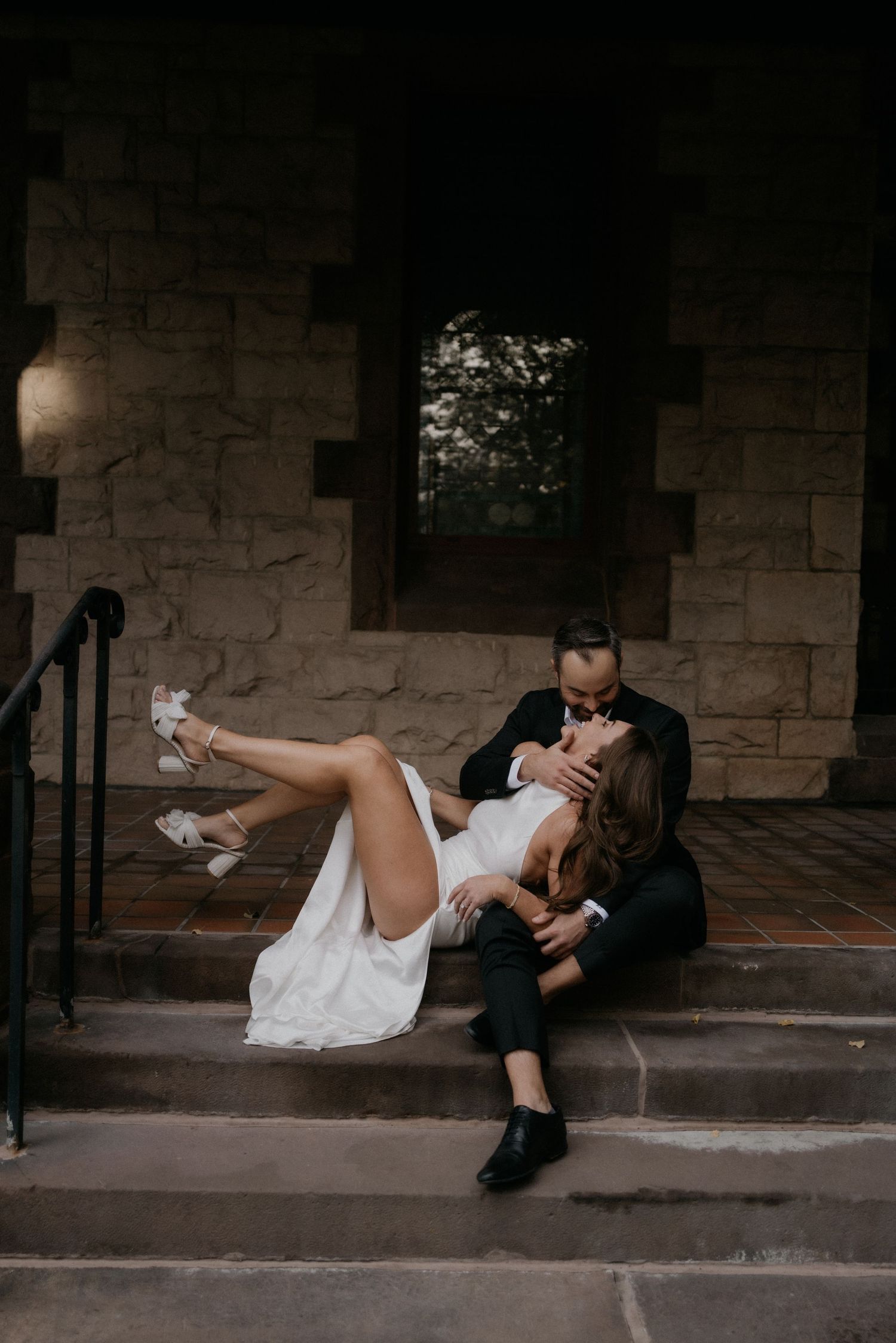 Couple in wedding attire poses dramatically sitting on ground against stone wall in moody lighting sequence.