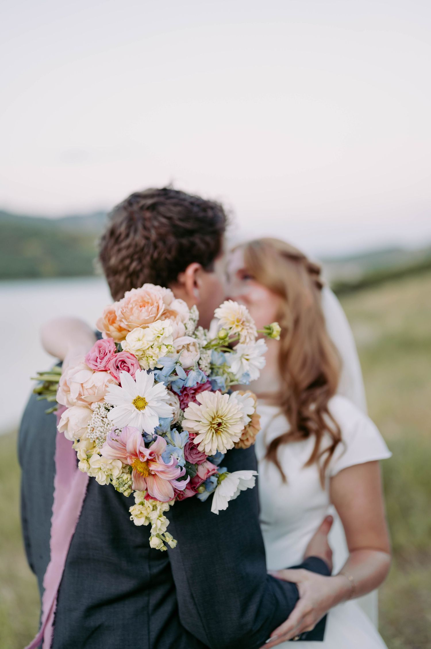 Wedding couple embracing with colorful pastel bouquet of flowers against scenic mountain backdrop.