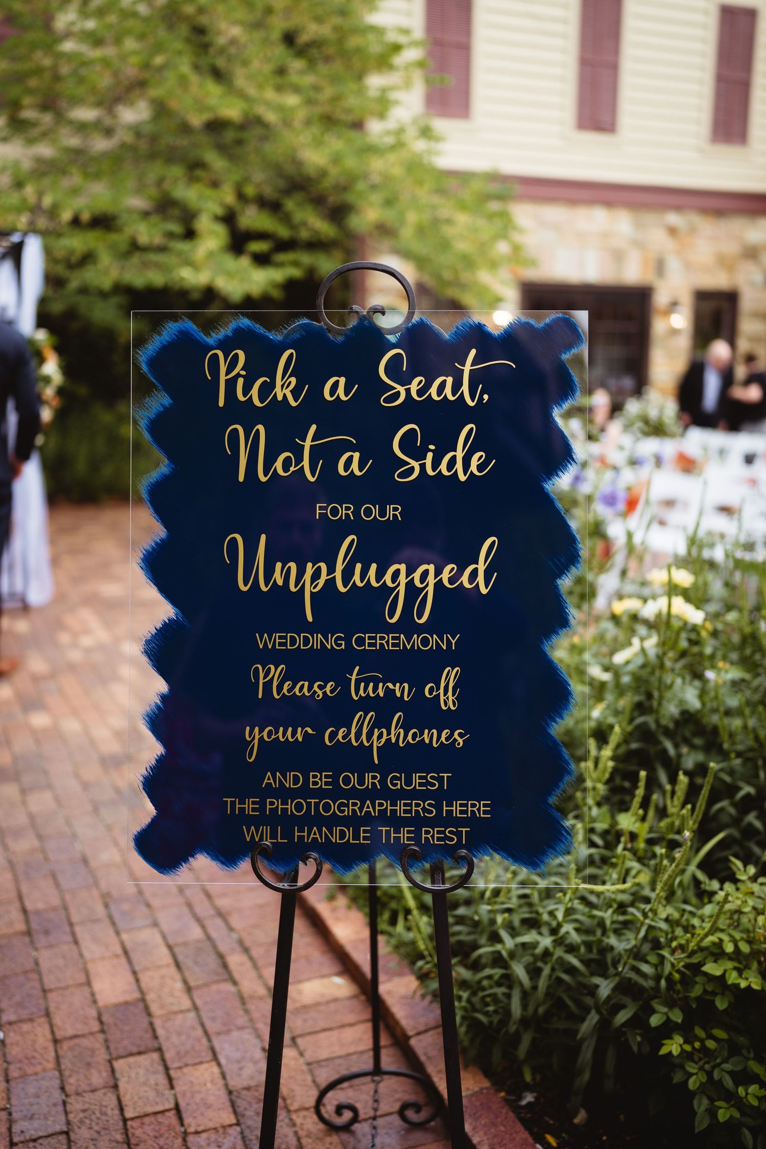 Three elegant wedding signs with gold script text on navy blue backgrounds displayed on decorative stands along a brick path.