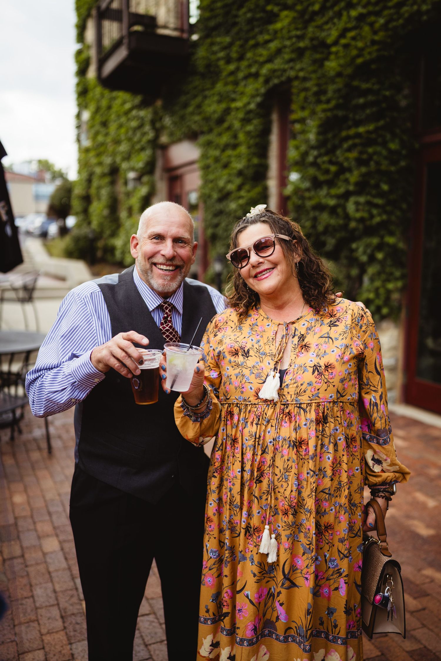 Two people share drinks and smile together on a brick sidewalk by an ivy-covered building wall.