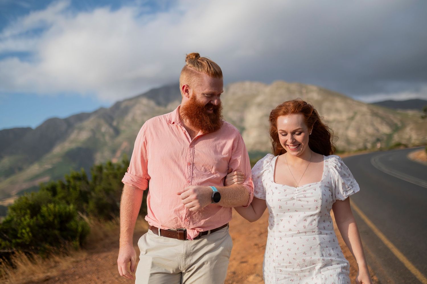 A couple in pink and white attire shares affectionate moments against a scenic mountain landscape backdrop during a photoshoot.