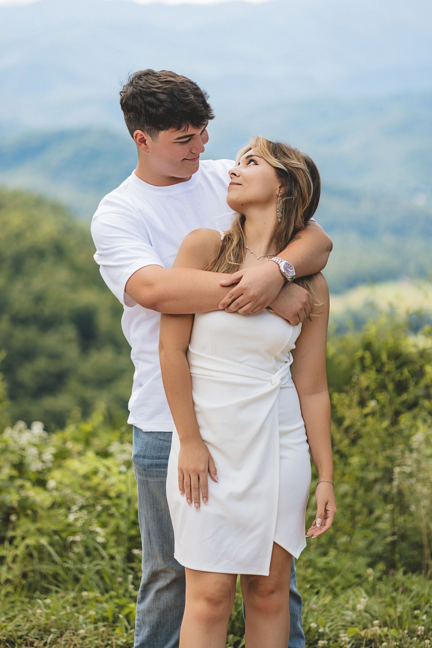 A romantic couple dressed in white embraces in a lush mountain setting with scenic views.