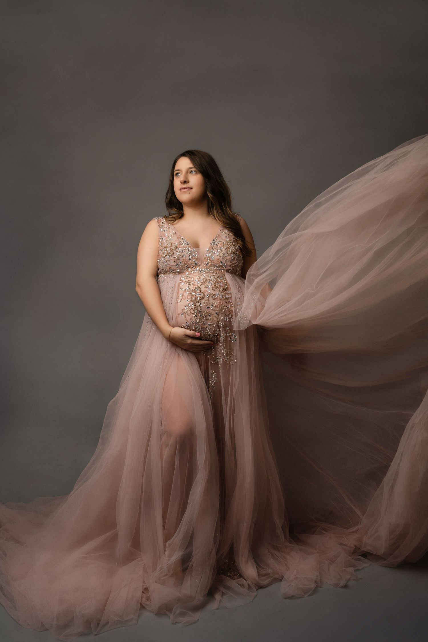 Pregnant woman in flowing blush tulle gown cradling her belly against a soft gray studio backdrop.