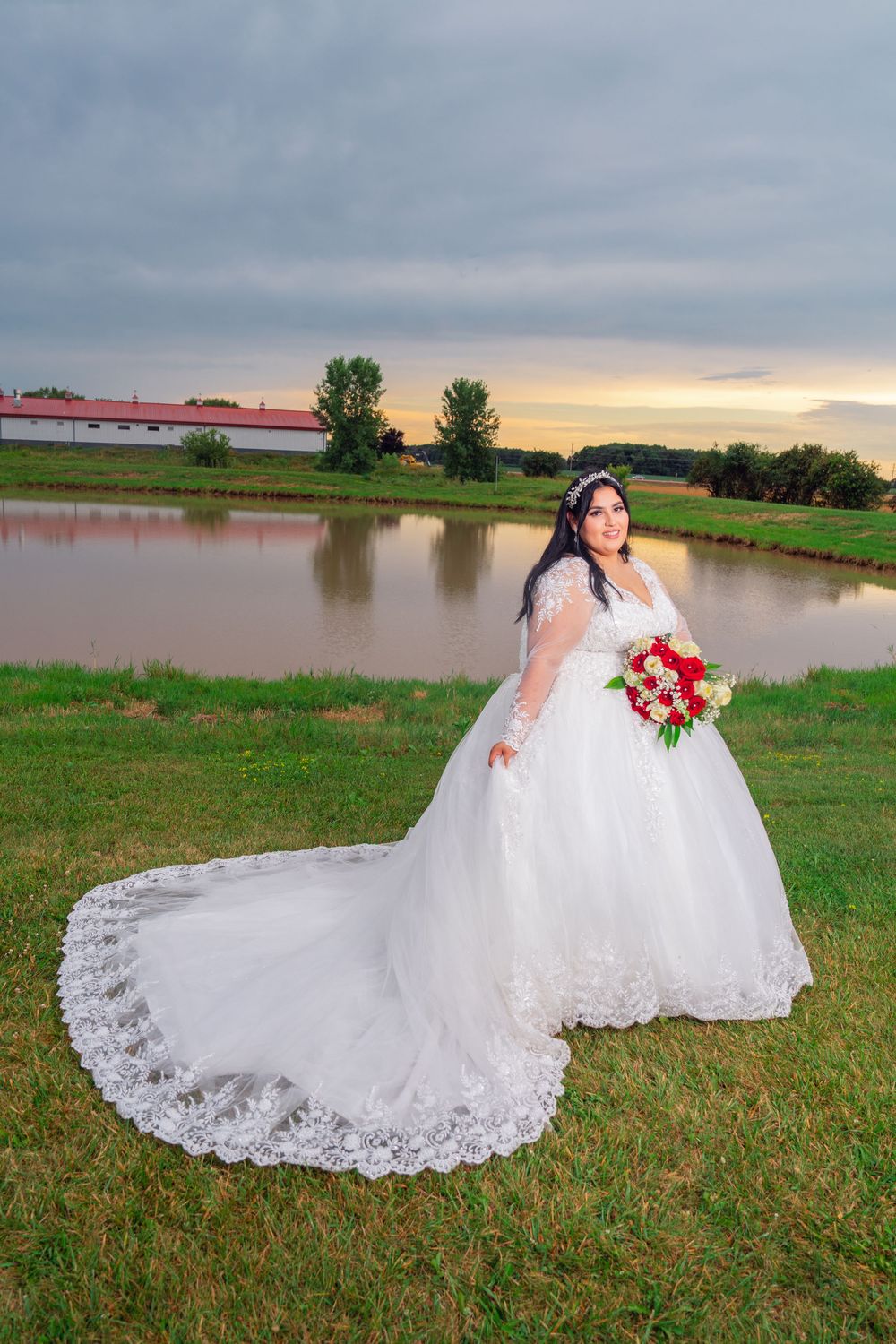 Elegant lace wedding dress with long train spreads across green grass near a lake. Appleton Photographer.