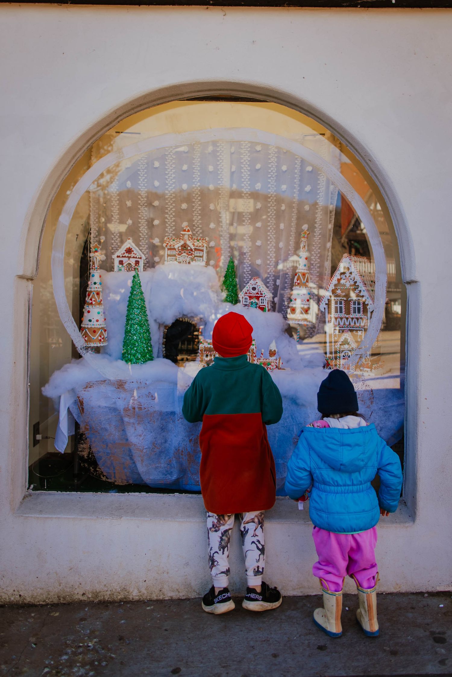 Two children look through an arched window display of Christmas decorations with twinkling lights and miniature trees.