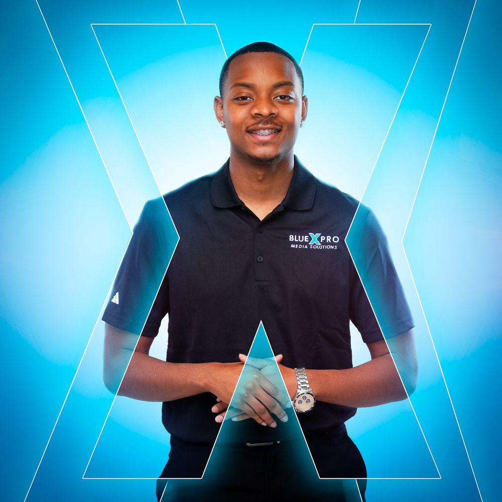 Professional staff member wearing a black polo shirt and slacks stands with clasped hands against gray background.