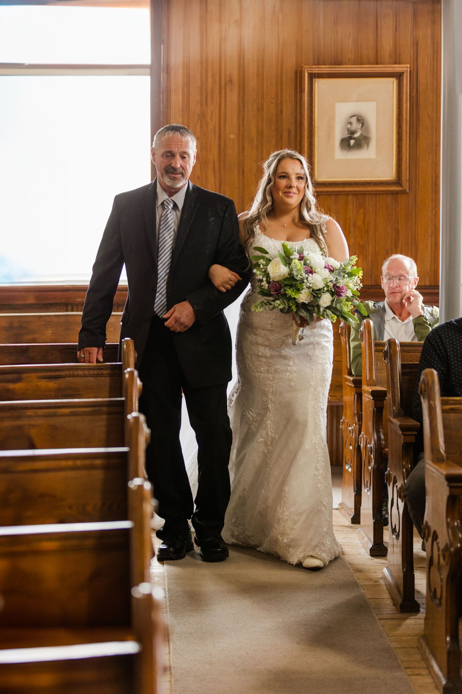 A wedding party progresses down a church aisle with wood paneled walls and pews in a touching processional moment.