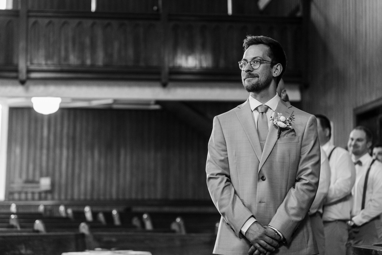 A groom in a light gray suit and bow tie waits expectantly at the altar during a wedding ceremony.
