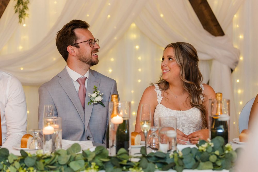 Wedding party seated at decorated head table with white drapery and string lights in background.